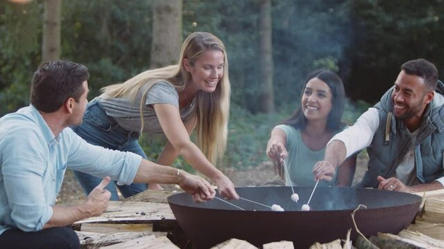 Group of friends on camping trip in forest sitting by fire toasting marshmallows together - shot in slow motion