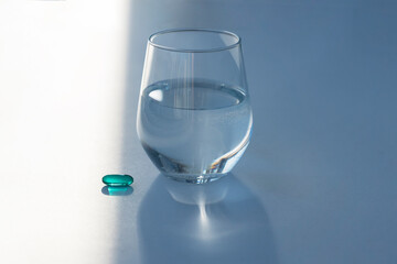 glass with pure water and  blue transparent pill capsule on white background, hard sunlight with shadows. Treatment and healthcare concept
