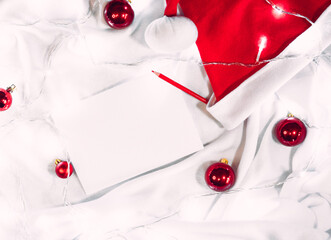 Creative Christmas banner and wish list sheet. A clean white sheet of paper and a red pencil lie on a blanket, and next to them are red Christmas balls, Santa's hat and bright lights of a garland.