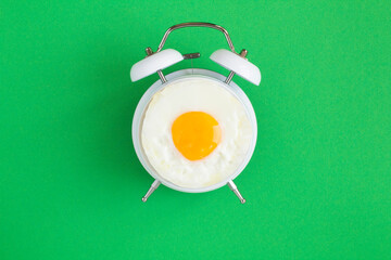 Top view of fried egg on the dial of the white alarm clock in the center of the green background