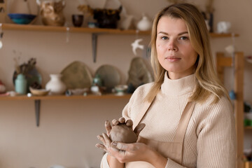 Serious caucasian beautiful blond female potter hold piece of clay in hands at a ceramic workshop place. Woman hobby and work concept