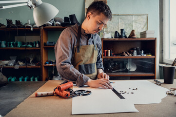 A young shoemaker makes a drawing for a pattern for leather shoes on a table in his workshop.