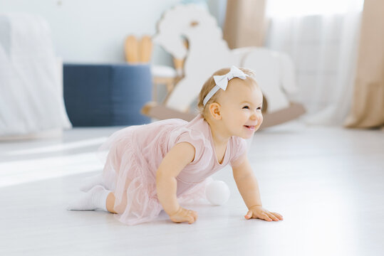 A pretty blue eyed blonde one year old in a pink dress crawls on all fours in the nursery on the floor and smiles fervently