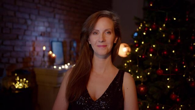 Smiling woman wishing Merry Christmas and Happy New Year. Portrait of woman in elegant black dress at home with Christmas tree.