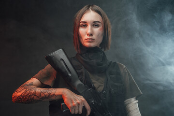 Seductive and tattoed woman dressed in modern and military body armour holding assault rifle in...
