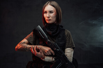 Seductive and serious female soldier with bandaged and tattooed hands poses in dark background holding rifle.