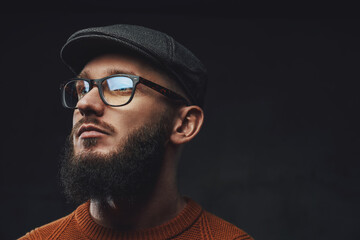Headshot of bearded hipster posing in dark background weared with glasses and cap.
