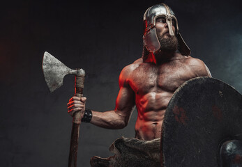 Muscular and bearded nord fighter with naked torso and helmet posing holding shield and axe in dark...