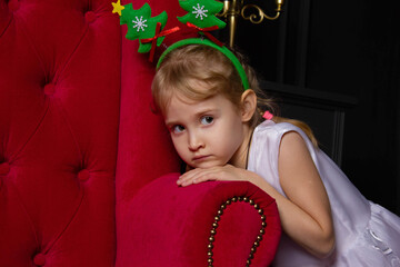 a pensive girl in a rim with Christmas trees leaned on the arm of a red chair, waiting for christmas