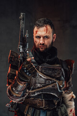 Holding pistol apocalyptic survivor in dark ragged armour and with damaged arm poses in dark background.