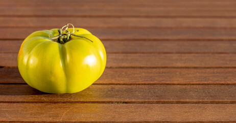 A single green raw village tomato on left side of frame. Natural food background on brown wood with copy space. Sun light reflecting on fruit.