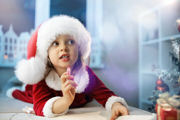 A cute little child girl dreams of writing a letter to Santa about a cherished gift, near the Christmas tree in her room