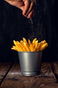 Hand salt delicious french fries in a metal cup. Dark background and old wooden table. Vertical frame.