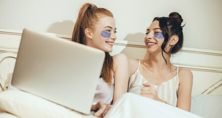 Caucasian friends with anti wrinkles masks under eyes laughing in bed with a laptop