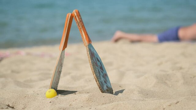 Two rackets and ball are on blue sea background. Matkot on the beach, blurred relaxing tourist. Beach tennis, paddle. Vacation on sea. Active healthy lifestyle. Catalonia, Spain.