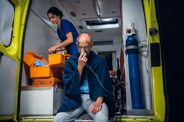 Man rescued from the fire sitting in an ambulance car, breathing through an oxygen mask, a female paramedic in the background. © Anna Kosolapova