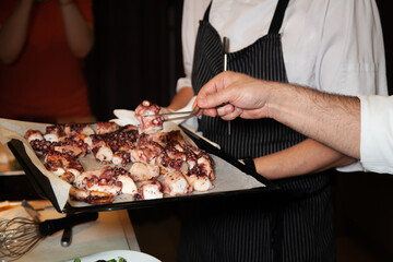 Two cooks having a grilled octopus on an oven tray, picking one bit to inspect it