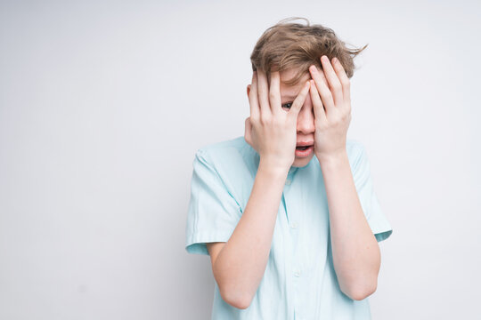 A Mid Shot Of A Frightened Caucasian Boy On A White Background. He Covered His Face With His Hands And Now He's Peeking Out Of His Fingers