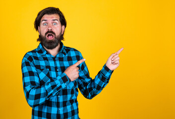 surprised handsome hipster in checkered shirt pointing away while standing against yellow...