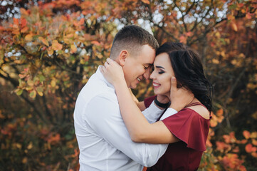 Fototapeta premium A stylish respectable guy in a white shirt and trousers and a beautiful girl in an expensive long dress stand and laugh, hugging against the background of red, yellow bushes with leaves.