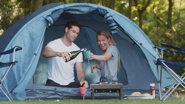 Couple in tent camping in countryside sitting by barbecue grilling opening bottle of champagne - shot in slow motion