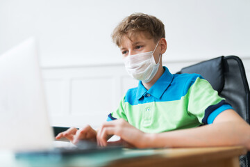 Fototapeta na wymiar Blonde American boy in a disposable face mask is actively participating in his online classes during the lockdown. He's typing the text on his laptop and listening to the teacher