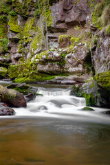 Fototapeta premium Small cascade on a mountain river flowing through the narrow, red rock canyon with rocks covered by green moss and cliffs with autumn colored trees