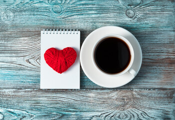 A red heart on a white Notepad and a Cup of coffee on a wooden gray-blue background. Top view with space to copy. Concept February 14.