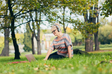 Senior woman resting in the park sitting on the grass, using phone and laptop, and reading a book