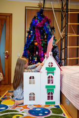 A little girl in a dress plays in a Dollhouse near a decorated Christmas tree waiting for Santa Claus