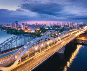 Aerial view of beautiful bridge at night in Kiev, Ukraine. Landscape with bridge, river, city illumination, blue sky with clouds at sunset. Cityscape with road, cars, buildings, city lights. Top view