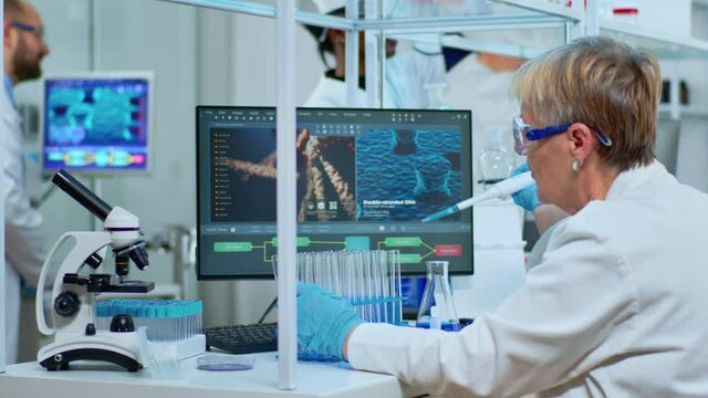 Elderly research scientist using micropipette filling test tubes in modern equipped lab. Multiethnic team examining virus evolution using high tech analysing treatment development against covid19.