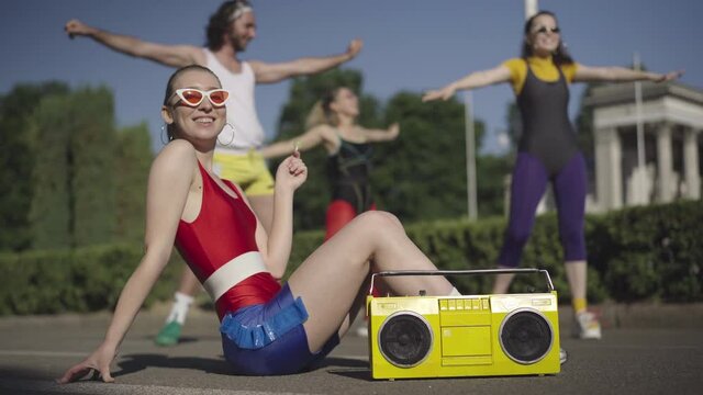 Confident gorgeous slim 1980s woman in sportswear and sunglasses sucking lollipop sitting with yellow vintage tape recorder as group of blurred people exercising at the background. Retro lifestyle.