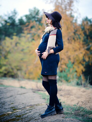A young beautiful pregnant woman in a hat and a black tight dress walks in the autumn nature.