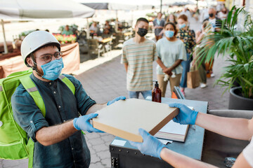 Delivery man wearing mask and protective gloves receiving pizza while collecting orders from the pickup point during coronavirus lockdown