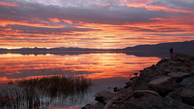 Silhouette of person taking photos of colorful sunset at Utah Lake reflecting the sky in the smooth surface of the water.