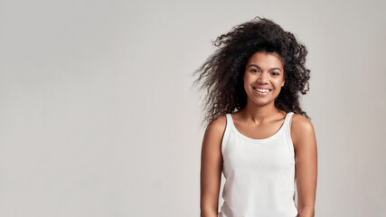 Portrait of young african american woman with curly hair wearing white shirt smiling at camera while standing isolated over grey background