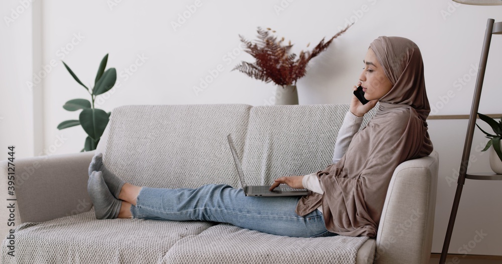 Wall mural modern work at home. side view shot of young islamic lady working on laptop and talking on phone