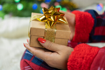 
a girl in santa claus pajamas holding a box of surprise gift with a gold bow for the new year holiday on the background of a Christmas tree with a garland