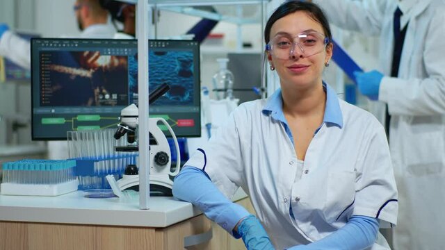 Portrait of scientist smiling at camera sitting in modern equipped laboratory. Multiethnic team examining virus evolution using high tech and chemistry tools for scientific research, vaccine