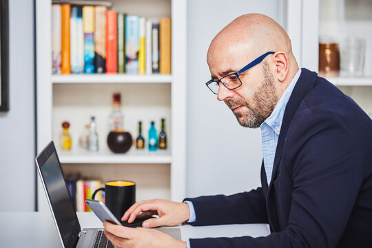 Business man teleworking from home with a laptop sending a message with his cell phone.
