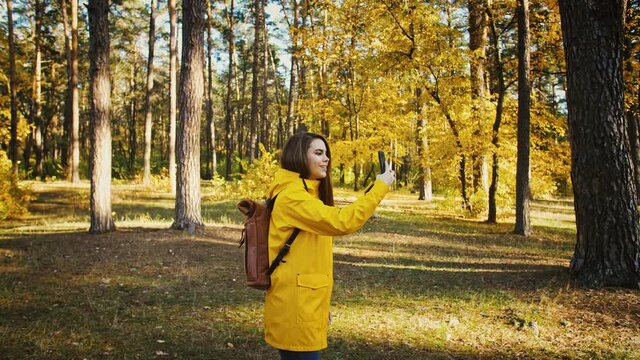 Young female smiling, taking photos of environment on her cellphone during walk in autumn forest