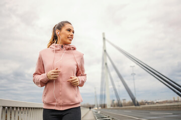 Fit sportswoman in tracksuit listening music and running on the bridge at cloudy weather. Urban exterior, healthy lifestyle concept.