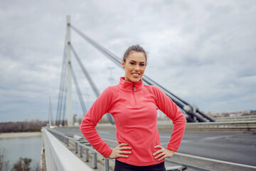 Smiling beautiful fit sportswoman standing on the bridge at cloudy autumn day with hands on hips...