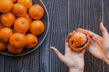 close up on a plate of ripe orange tangerines on a wooden table, one tangerine peeled in woman's hands - 395141821