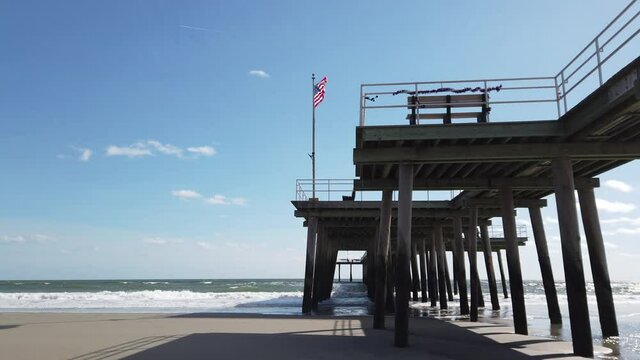 View from under a long wooden pier showing the beach and ocean waves hitting the pilons. There is an American Flag on the pier waving in the wind