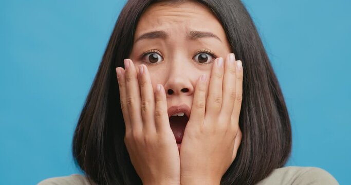 Close Up Of Shocked Amazed Asian Woman Touching Face In Astonishment And Wonder, Blue Studio Background