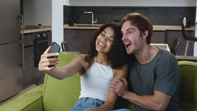 Multiracial couple relaxing on couch taking selfie pictures using smartphone, making face at camera, having fun together posing for selfie on mobile phone for social media.