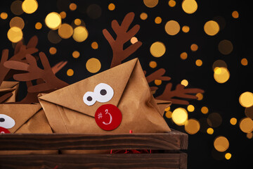 Gifts in envelopes with deer faces in wooden crate against blurred lights, closeup. Christmas advent calendar