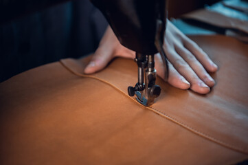 Close-up of the sewing machine needle guide makes an even seam with a thread on a brown cloth of genuine leather. The hands of the cobbler.